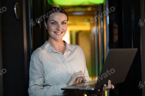 Preview: Portrait of caucasian female engineer smiling while using laptop in computer server room