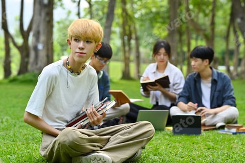 Preview: Asian male student sitting on green grass in campus with friends sitting on background.