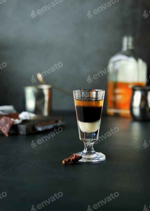 Preview: A three-layer cocktail shot with whiskey and liqueurs in a glass and chocolate next to it on a dark