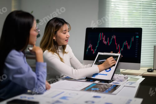 Preview: Businesswomen analyzing financial charts and graphs on computer and clipboard in office