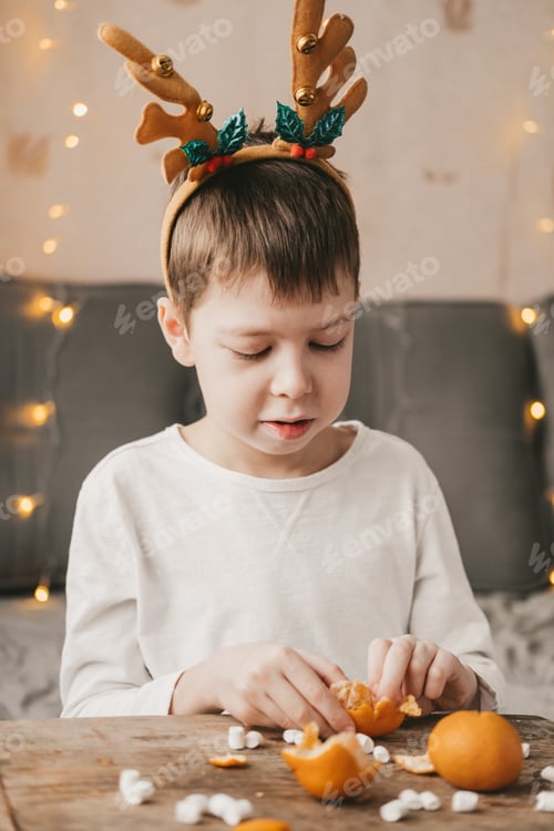 Preview: boy in antlers,peels a tangerine of a garland. Boy in a white t-shirt at home, peels a tangerine