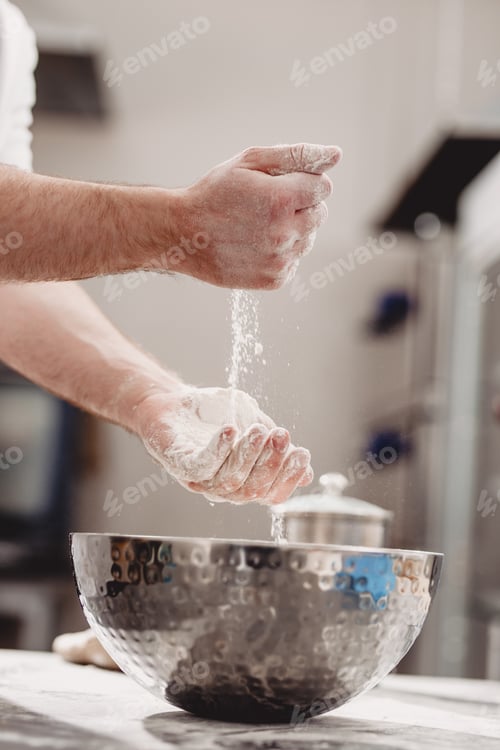 Preview: Baker adds flour to dough in an iron bowl on the table in the bakery