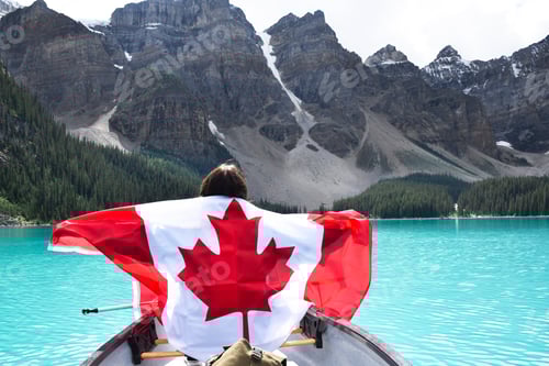 Preview: Young girl from behind with Canadian flag riding in canoe on turquoise lake surrounded by mountains