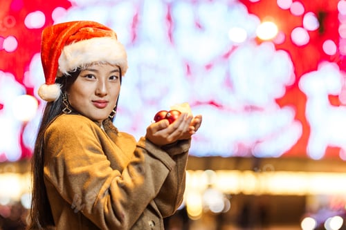 Preview: Young woman wearing santa hat holding christmas baubles in city at night