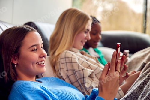 Preview: Group Of Multi-Cultural Teenage Girl Friends Snuggled Under Blanket Looking At Mobile Phones At Home