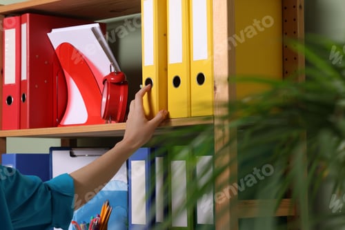 Preview: Woman taking binder office folder from shelving unit indoors, closeup