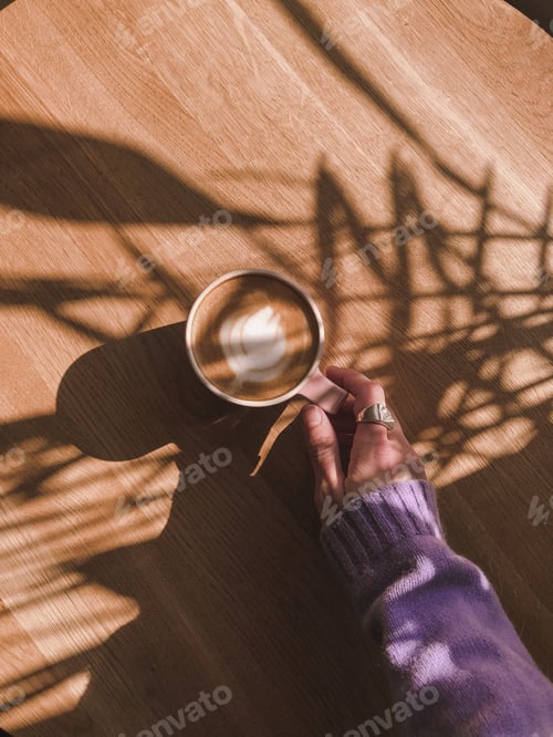 Preview: Female hand holding cup of coffee on wooden background with beautiful shadows of the leaves.
