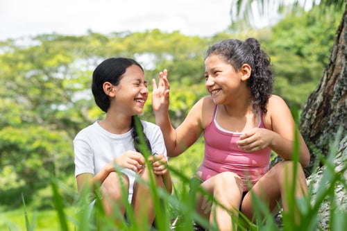 Preview: two brunette latina girls, sitting next to a tree in a park playing and laughing together