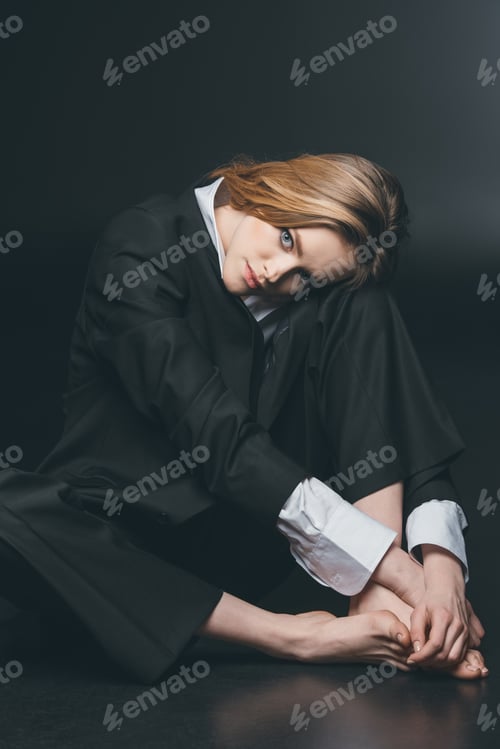 Preview: Young adult woman sitting and posing in male costume on black background, studio shot