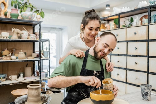 Preview: Smiling craftswoman in apron hugging boyfriend coloring clay bowl in ceramic workshop at background
