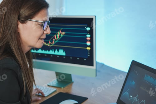 Preview: Mature trader woman working at home with computer and laptop doing stock market research