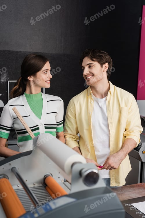Preview: cheerful woman holding ruler and looking at happy colleague near print plotter machine