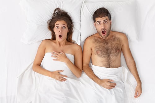 Preview: Overhead shot of emotional shocked woman and man stay in bed covered by sheet, stare with wide opene
