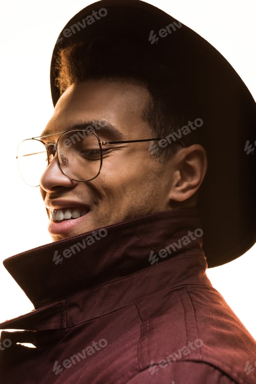 Preview: stylish mixed race man in glasses, hat and coat smiling and posing isolated on white