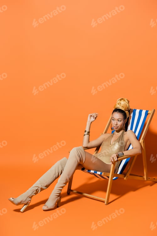 Preview: Full length of stylish african american woman in swimsuit and headscarf sitting on deck chair on