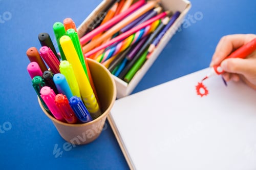 Preview: Child's hands drawing with colorful pencils and markers in a notebook on blue background.
