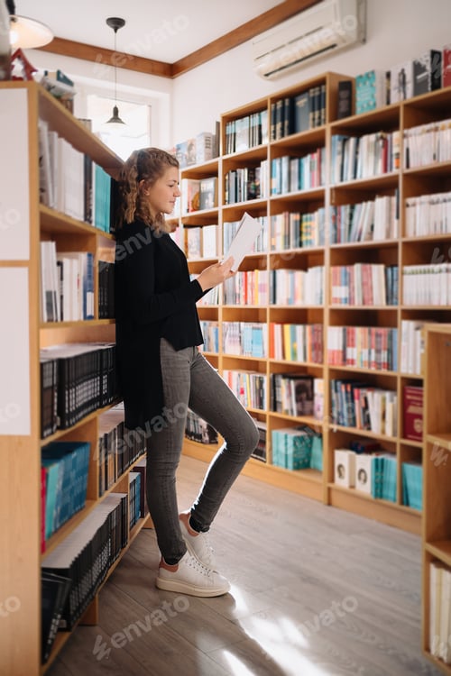 Preview: Beautiful girl is studying reading a book while standing on the floor among books in the bookshop