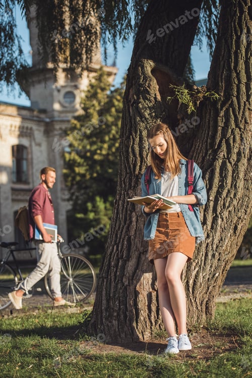 Preview: young student girl reading homework and leaning back on tree in park