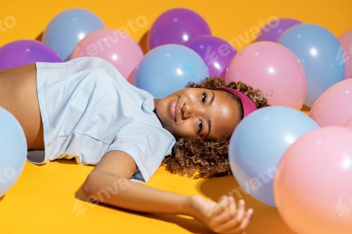 Preview: Young woman lying on yellow background surrounded by colorful balloons