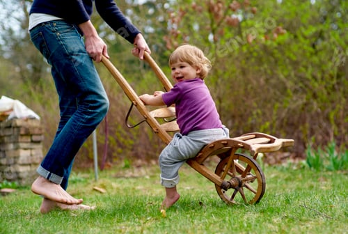 Preview: Toddler enjoys a ride in a wooden wagon outdoors, pulled by an adult on a sunny day.