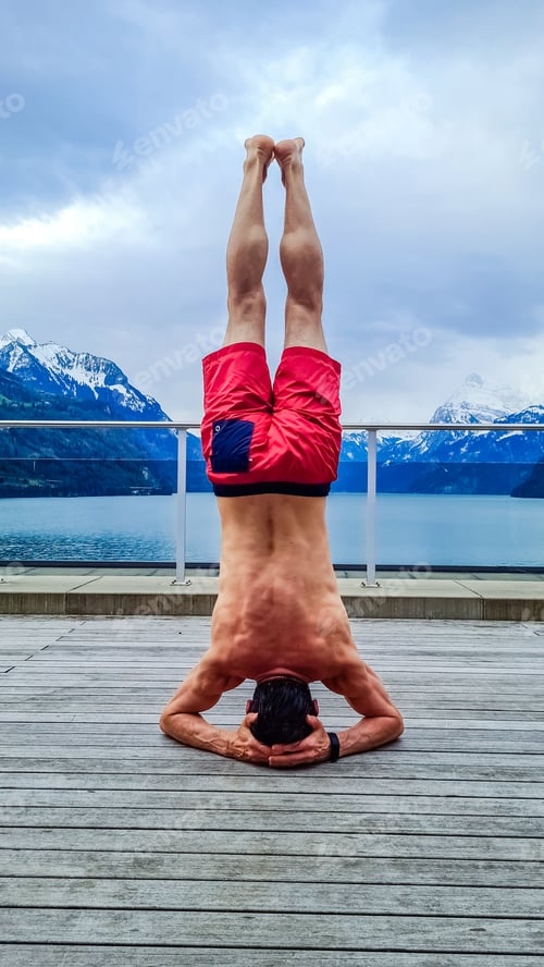 Preview: Healthy lifestyle. Young fit man doing head standing exercise outdoors with beautiful mountain view
