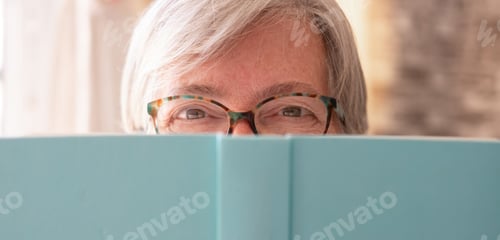 Preview: Portrait of beautiful caucasian senior woman partially hidden by a book looking at camera smiling