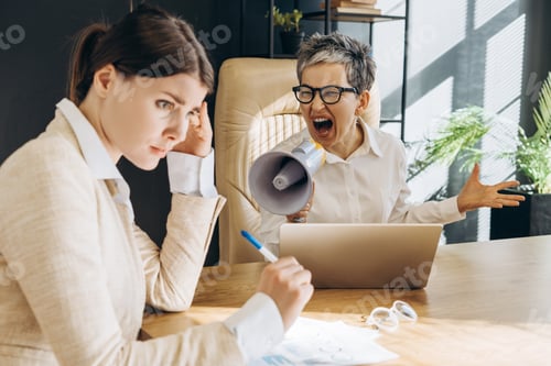 Preview: Angry senior manager shouting at young employee through megaphone in office