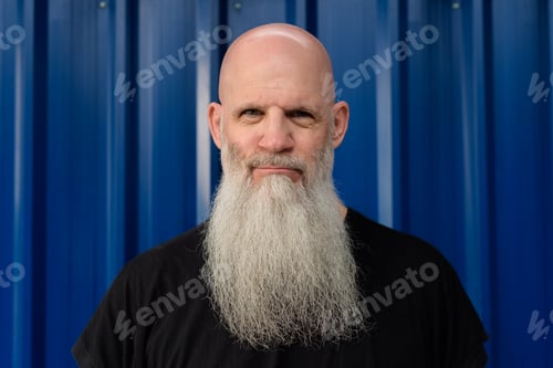 Preview: Mature handsome bald bearded man looking at camera against metal sheet wall