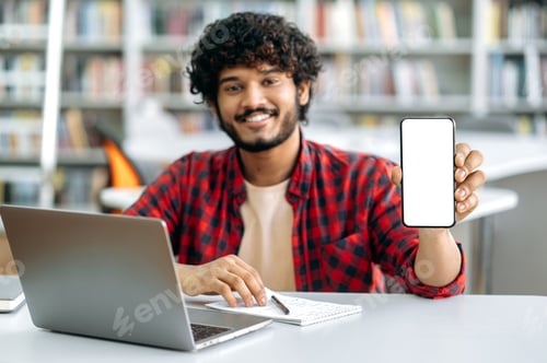 Preview: Defocused Indian or Arabian male student, sitting at table in library, showing his smart phone with