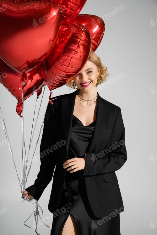 Preview: happy young woman in black slip dress and blazer holding red heart-shaped balloons isolated on grey