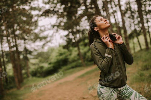 Preview: Young beautiful female runner listening to music and taking a break after jogging in a forest