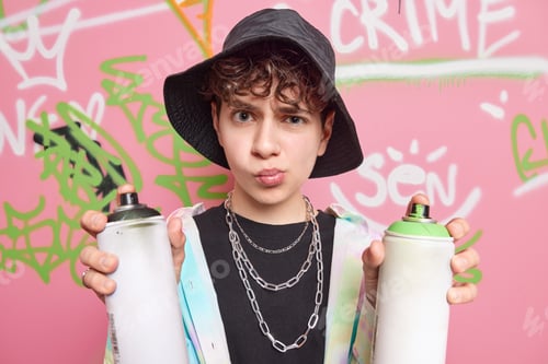 Preview: Discontent urban street artist wears fashionable clothes holds two aerosol sprays smirks face looks