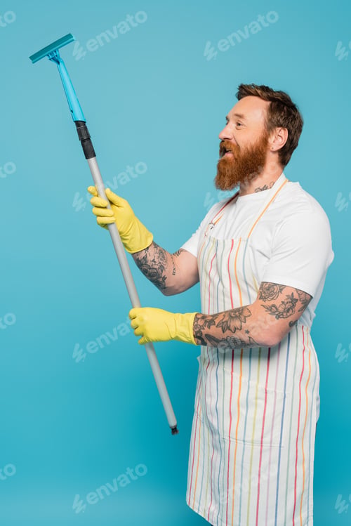 Preview: smiling tattooed man in striped apron and yellow rubber gloves holding window scraper isolated on