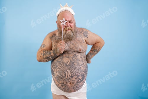Preview: Tattooed obese man with crown looks through star on magic stick on blue background