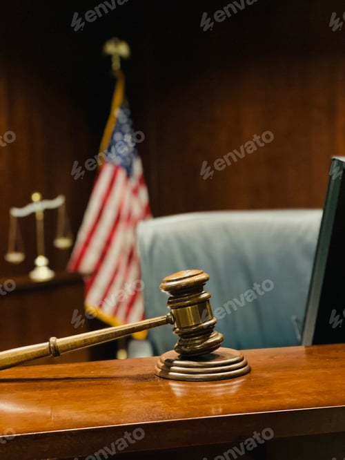 Preview: Selective focus on gavel, judge’s bench, scales of justice and flag in background.