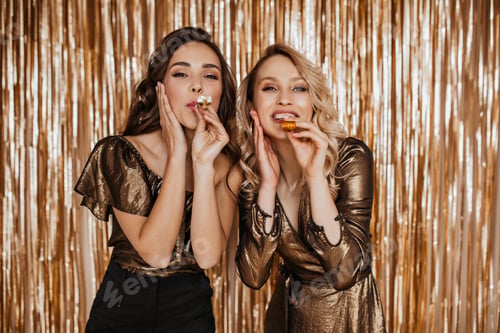 Preview: Brunette girl and blonde girl wearing similar outfit are blowing in festive pipes against gold back