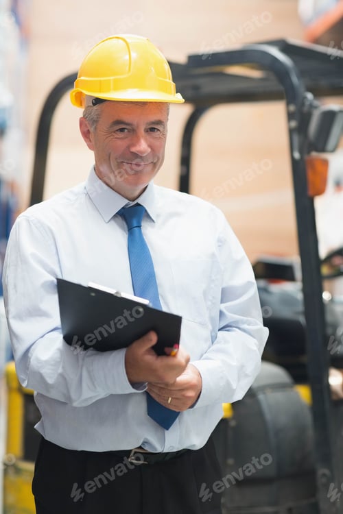 Preview: Male supervisor wearing yellow helmet holding clipboard inspecting goods near forklift in warehouse