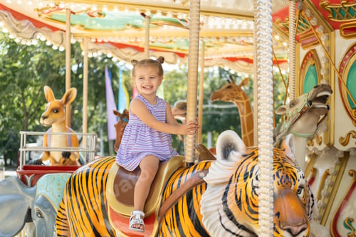 Preview: funny little kid girl in colorful dress rides on carousel in an amusement park in summer day