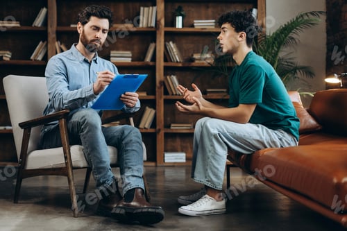 Preview: Teenager boy sitting on couch while therapist listening and taking notes