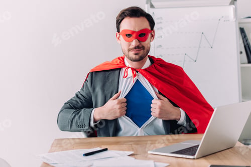 Preview: handsome super businessman in mask and cape showing blue shirt in office