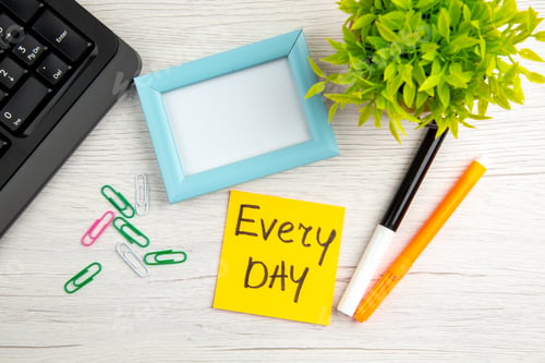 Preview: Top view of blue empty picture frame and every day note half shot laptop paper clips marker flower