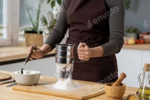 Preview: Hands of young woman in apron sieving flour for homemade pastry
