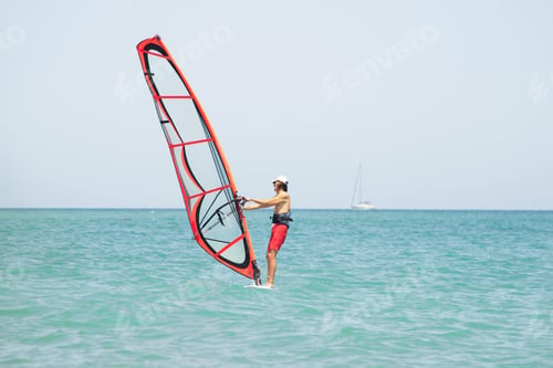 Preview: a man controls a sail on a surfboard in the sea against the background of the water and the sailboat