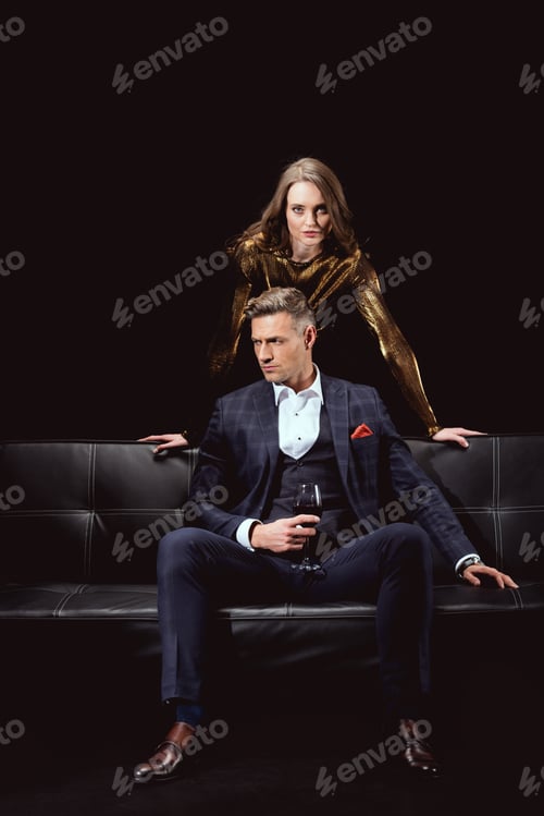 Preview: handsome man in formal wear holding glass of red wine while woman behind looking at camera isolated