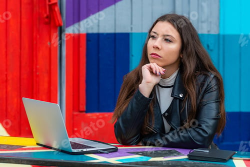 Preview: thoughtful young woman with laptop sitting at table in outdoor cafe