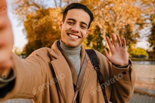 Preview: Young man wearing coat waving hand and taking selfie photo in park