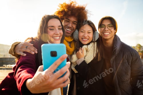 Vista previa: Mujer joven que se toma una selfie con amigos multirraciales al aire libre en un día soleado de invierno con un teléfono móvil.