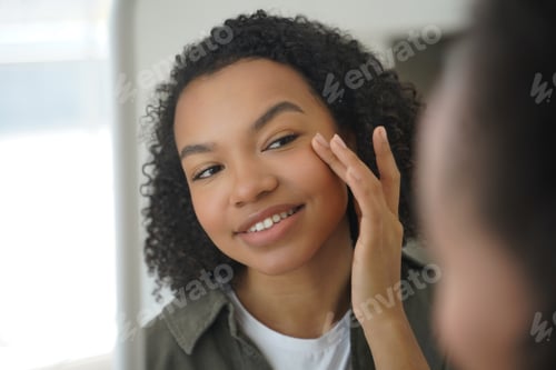 Preview: Biracial young girl looks at mirror, applying moisturizing facial cream. Natural beauty, skincare
