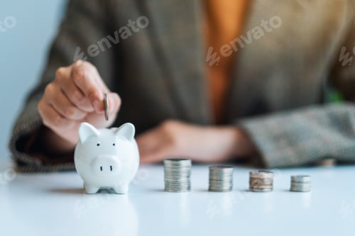 Preview: A businesswoman putting coin into piggy bank with coins stack on the table for saving money