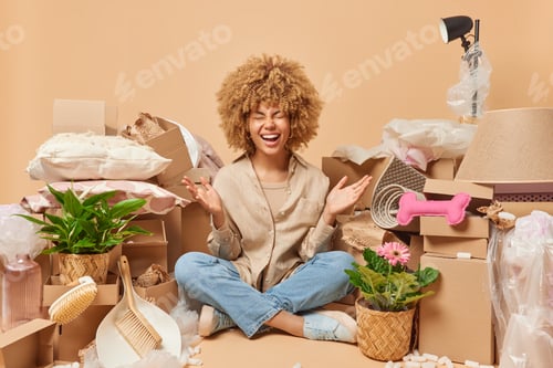 Preview: Happy young woman wears shirt jeans and sportshoes sits in lotus pose on floor in messy room relocat